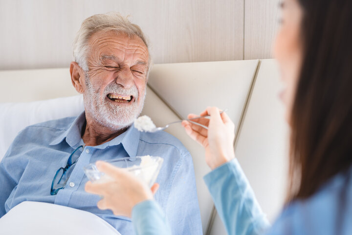 Elderly patient having support health care service by nurse in a old people home providing residential care by bringing breakfast to her senior man occupant, medical care of retirement man