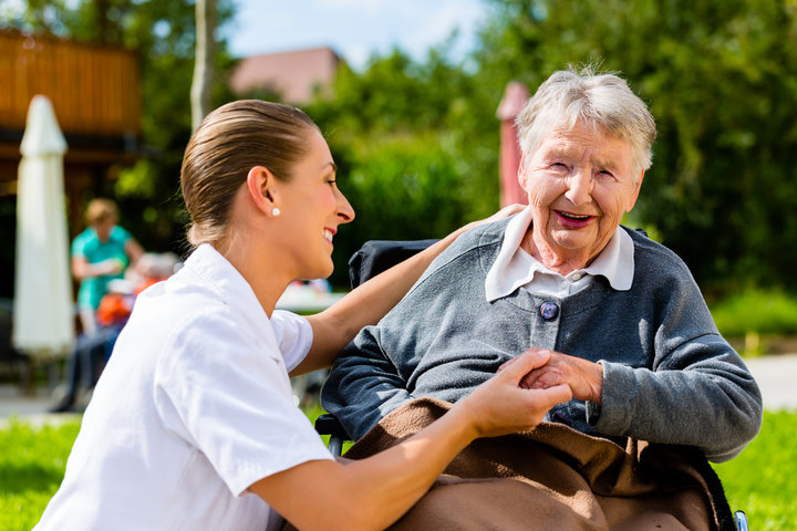 older woman in a wheelchair outside in a garden with a nurse bending down helping her